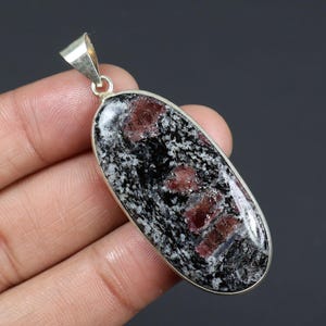 Puede incluir: Colgante ovalado con una fianza plateada. El colgante presenta una piedra moteada en blanco y negro con parches de color marrón rojizo. La piedra está engastada en un bisel plateado. El colgante está sostenido por una mano.