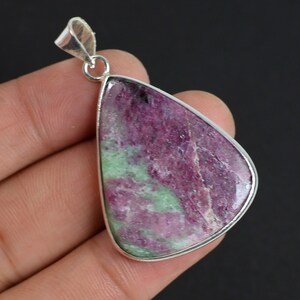 May include: A pendant with a large, irregularly shaped ruby zoisite stone. The stone is mainly a deep pink with green inclusions, set in a silver-coloured metal frame. The pendant has a loop at the top for a chain.