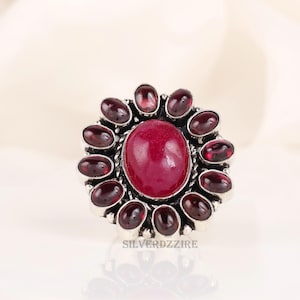 Op de afbeelding: Een zilveren ring met een grote, ovale, dieprode edelsteen in het midden, omringd door kleinere, ovale, donkerrode edelstenen. De ring heeft een gedetailleerde, getextureerde zetting. De tekst "SILVERDZZIRE" is zichtbaar op de ring.