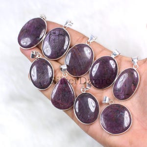 May include: A selection of oval and teardrop pendants featuring deep purple gemstones set in silver-coloured metal. The pendants are held in a hand, highlighting their polished surfaces and unique designs. The gemstones have a natural, textured appearance.