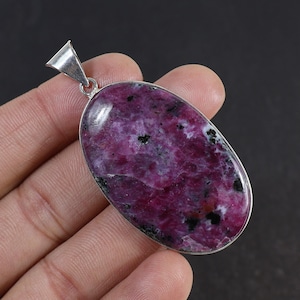 Puede incluir: Un colgante de forma ovalada con una piedra pulida de color magenta intenso y púrpura con inclusiones negras. La piedra está engastada en un bisel de metal plateado liso con una anilla triangular en la parte superior para una cadena.