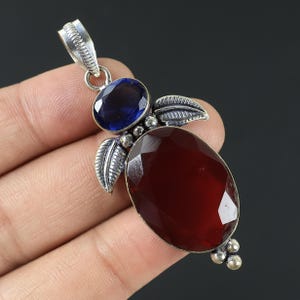May include: A silver pendant featuring a large, faceted, oval-shaped, deep red gemstone and a smaller, oval, dark blue gemstone. The pendant has leaf-shaped accents and a loop for a chain.