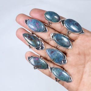 Op de afbeelding: Een verzameling edelsteenringen met ovale en druppelvormige stenen. De stenen zijn een mix van blauwgroen met rode insluitsels, gezet in zilverkleurig metaal. De ringen worden op een hand getoond.