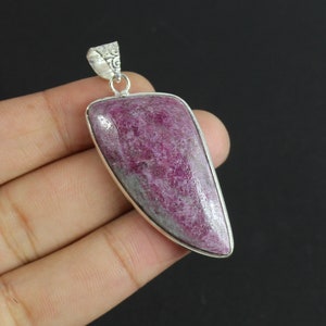 May include: A teardrop-shaped pendant featuring a textured, deep magenta and grey stone set in a polished silver-toned metal bezel. The pendant includes a decorative, etched silver-toned bail at the top for attachment to a chain or cord.