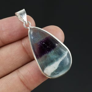 Può includere: Un ciondolo a forma di goccia con una pietra preziosa di fluorite lucida caratterizzata da bande di viola intenso, verde acqua traslucido e bianco opaco, incastonato in una montatura in metallo color argento con una contromaglia triangolare.
