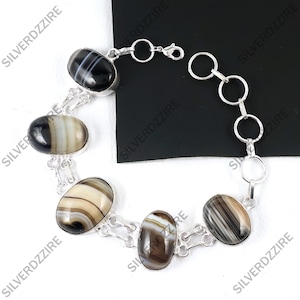 Puede incluir: Un brazalete de plata con piedras de ágata ovaladas y bandeada en tonos negro, marrón y crema. Las piedras están engarzadas en una cadena de plata con un cierre de mosquetón. El brazalete se exhibe sobre una superficie blanca.