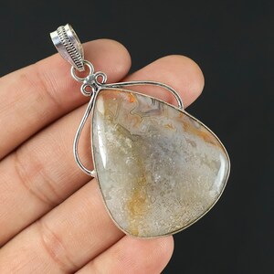 Puede incluir: Un colgante en forma de lágrima que presenta una piedra de ágata pulida multicolor con patrones de remolinos en blanco, gris y ámbar, engastada en un marco decorativo de plata esterlina con una anilla texturizada en la parte superior.