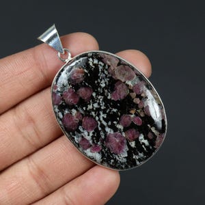Peut inclure: Pendentif ovale avec bélière argentée. Le pendentif présente un fond moucheté noir et blanc avec des pierres de couleur rubis incrustées. Le pendentif est tenu dans une main sur fond sombre.