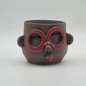 May include: A dark brown ceramic mug shaped like a face with large red circular glasses, a small nose, and red lips. The mug features small ear-like protrusions on the sides, each adorned with a tiny heart-shaped detail, creating a stylized character.