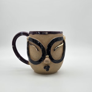 May include: A ceramic mug with a face design. The mug features black-rimmed glasses, closed eyes, and a small mouth. The handle is dark purple with white polka dots, and the rim is dark purple with small white dots.