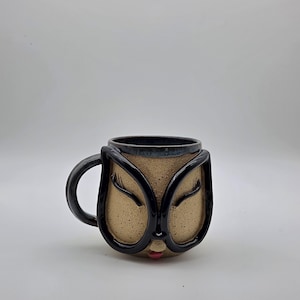 May include: A ceramic mug with a speckled beige finish featuring a sculpted face design. The mug has black raised outlines forming large glasses around closed eyes with long eyelashes, and a small red painted mouth at the bottom center.