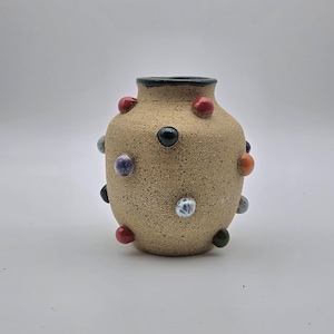 May include: A small, round, speckled tan ceramic vase with a black rim. The surface is decorated with several raised, smooth, multi-colored spheres in shades of red, black, orange, white, and purple, creating a textured and tactile appearance.