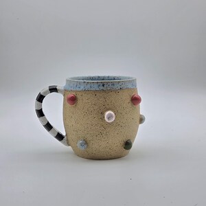 May include: A ceramic mug with a speckled tan body, a light blue speckled rim, and a black and white striped handle. The exterior features several small, raised, multi-colored circular dots in shades of pink, red, blue, and dark green.