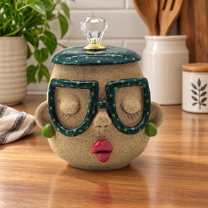 May include: A ceramic jar shaped like a face with a speckled tan finish, wearing large dark green glasses with light green dots. The lid features a clear crystal-like knob, and the face has bright pink lips, closed eyes, and small green earrings.