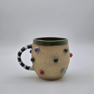 May include: A speckled beige ceramic mug with a dark green rim and a black and white striped handle. The exterior surface is decorated with small, raised, multi-colored circular dots in shades of red, blue, black, and purple, creating a textured look.