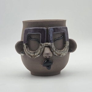 May include: A brown ceramic mug shaped like a human head with closed eyes, a nose, and ears. The mug features large, dark purple and textured grey glasses over the eyes, and glossy black lips. The surface of the mug has a matte, earthy finish.