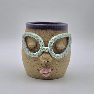 May include: A speckled tan ceramic mug featuring a sculpted face with closed eyes, a nose, and pink lips. The mug has a dark purple rim and is adorned with light blue, green, and purple speckled glasses resting over the eyes of the face.