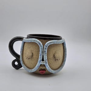 May include: A ceramic mug with a speckled tan finish featuring a sculpted face with closed eyes, red lips, and large light blue glasses. The mug has a dark brown interior and a dark brown handle that curves outward from the side of the vessel.