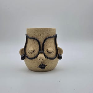 May include: A speckled beige ceramic planter shaped like a face with closed eyes, a small nose, and black lips. The face features prominent black glasses frames and small black earrings on each side, creating a distinct, sculptural appearance.