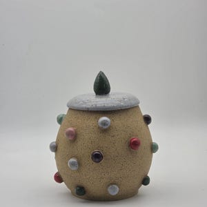 May include: A round, speckled tan ceramic jar with a removable lid. The lid features a dark green, pointed handle. The jar is decorated with various small, raised, colorful spheres in shades of red, blue, pink, and dark green across its surface.