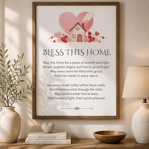 Op de afbeelding: Een ingelijste print met de woorden "BLESS THIS HOME" en een gedicht over thuis. Het kunstwerk toont een aquarel illustratie van een huis, harten en een groot roze hart. De lijst is bruin en de print wordt op een houten tafel getoond.