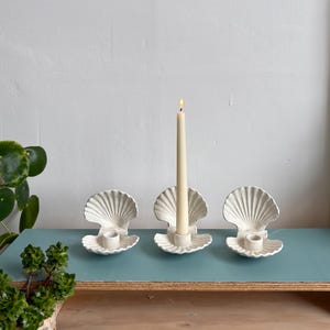 May include: Three off-white ceramic candle holders shaped like open scallop shells. Each holder features a central cylindrical socket for a taper candle. One tall, slender, cream-coloured taper candle is lit and placed in the centre holder.
