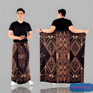 Puede incluir: Un sarong negro con un patrón geométrico dorado y marrón. El sarong está doblado y drapeado sobre el brazo de una persona.