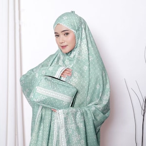 May include: A light green prayer shawl with a white floral pattern and lace trim. The shawl is folded and held by a person. A small, rectangular pouch with a white lace trim is also visible.