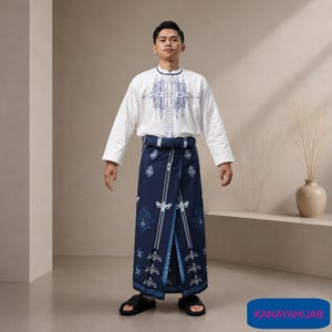 May include: A white long-sleeved shirt with a blue geometric chest pattern and a dark blue sarong featuring white floral and linear motifs. The outfit is paired with black sandals. The text KANAYAHIJAB appears in a blue box in the corner.