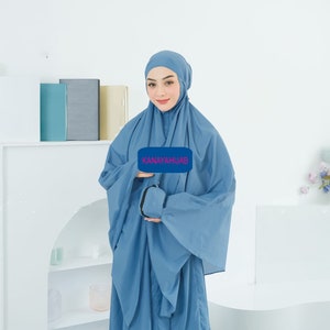 May include: A woman wearing a light blue prayer dress and matching headscarf. The dress has a pocket on the front and the text "KANAYAHIJAB" is printed on a white rectangle.