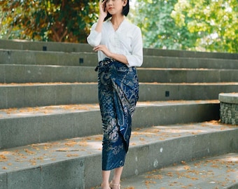 May include: A woman wearing a white long-sleeved shirt and a blue and gold patterned wrap skirt. She is standing on a set of concrete stairs.