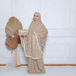 May include: A beige prayer outfit with a brown leaf pattern and white lace trim. The outfit includes a long skirt and a matching top with a hood. A small beige bag is included.