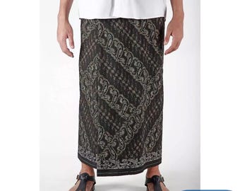 May include: A black and gray sarong with a paisley pattern. The sarong is worn by a person. The bottom of the sarong has a black border. The person is wearing black sandals. The text "KANAYAHIJAB" is in a blue rectangle.