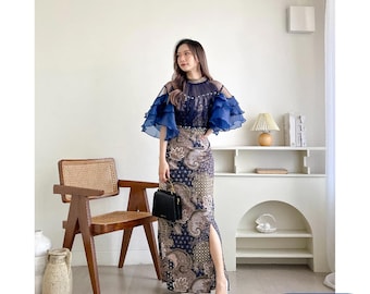 May include: A woman wearing a long, blue and white patterned dress with a blue sheer overlay. The dress has a high neckline and a slit up the side. She is wearing white heels and holding a black handbag.