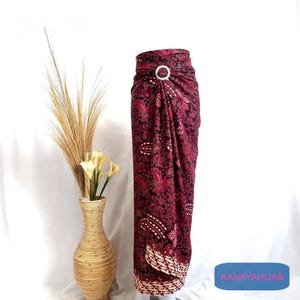 Op de afbeelding: Een sarong met een marroon en zwart patroon met een witte ringaccent. De sarong is gedrapeerd en geknoopt in de taille. De sarong is op een witte achtergrond.