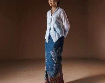 May include: A woman wearing a white blouse with blue watercolor accents and a long, dark blue skirt with a floral pattern. The skirt has a brown and orange border. The text "KANAYAHIJAB" is in a blue rectangle.