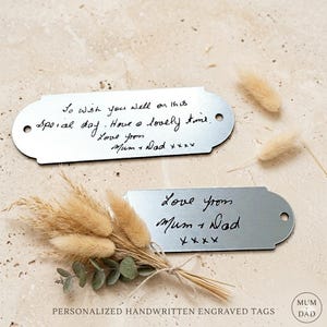 May include: Two silver metal tags with handwritten messages. One says "To wish you well on this special day. Have a lovely time. Love from Mum + Dad xxxx." The other says "Love from Mum + Dad xxxx." Dried flowers are also in the image.