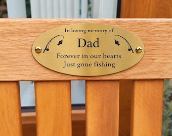 May include: An oval brass memorial plaque attached to a wooden surface with two screws. The plaque features black engraved text reading In loving memory of Dad, Forever in our hearts, Just gone fishing, and two black fishing rod icons.