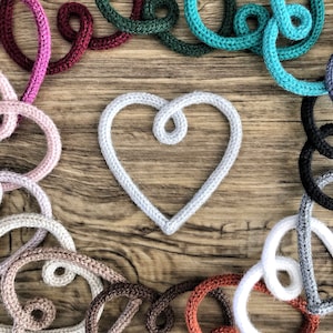 May include: A white crocheted heart shape surrounded by a variety of colourful crocheted loops on a wooden background.