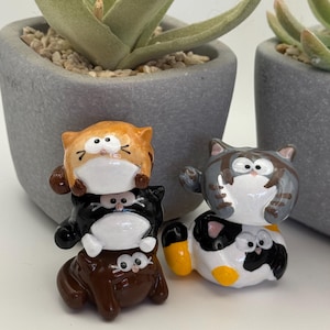 Puede incluir: Dos conjuntos de figuras de gatos. Uno está apilado en marrón, negro y naranja. El otro es gris, blanco y amarillo. Las figuras tienen caras blancas. El fondo incluye dos macetas grises con plantas verdes.