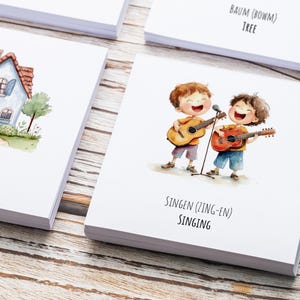 May include: Two children with brown hair are singing and playing acoustic guitars. The text on the card reads "SINGEN (ZING-EN) SINGING".