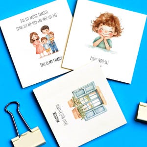 May include: Three illustrated cards with watercolor designs. The first card shows a family of four with the text "Das ist meine Familie (Dahs ist my-neh fah-mee-lee-eh) This is my family". The second card shows a girl with the text "Ruht (Roo-ig)". The third card shows a window with the text "Fenster (Fen-ster) Window".