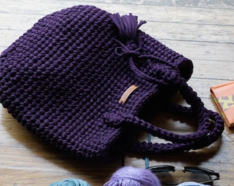 May include: A deep purple crocheted tote bag with a long tassel and a leather tag. The bag is lying on a wooden floor.