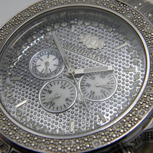 May include: A silver watch with a diamond-encrusted bezel and face. The watch has three sub-dials, one of which displays the days of the week. The watch face also features the word "LUXURMAN".