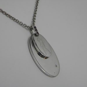 May include: A silver necklace with a double oval pendant. The top oval features a cutout of a woman's silhouette. The bottom oval is plain. The necklace has a row of small purple gemstones.