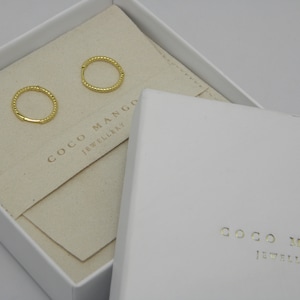 Könnte beinhalten: Zwei goldene Creolen in einer weißen Box mit dem Text "Coco Mango Jewellery" auf der Innenseite der Box.