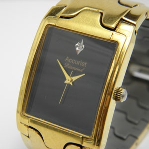 Könnte beinhalten: Goldfarbene Accurist Diamond-Uhr mit rechteckigem, schwarzem Zifferblatt. Die Uhr hat einen Diamantakzent und goldene Zeiger. Das Armband ist ebenfalls goldfarben. Klassisches Design.