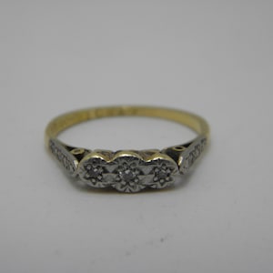 Könnte beinhalten: Ein gold-silberner Ring mit drei kleinen Diamanten in einer Reihe. Der Ring hat ein filigranes Design mit aufwendigen Details am Band und um die Diamanten. Ein klassisches Schmuckstück.