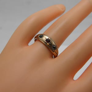 May include: A gold-toned band ring featuring three small, dark, round-cut stones set in a row. The metal surface of the ring is decorated with a subtle, engraved diamond-shaped pattern and a beaded border along the edges of the band.