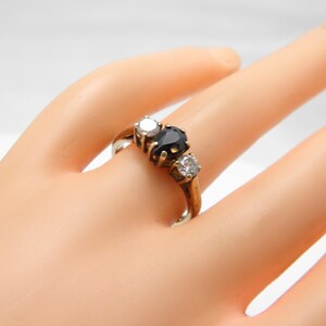 May include: A gold ring featuring a central pear-shaped dark blue gemstone flanked by two round clear stones. The band is polished gold, and the stones are set in a prong setting, creating a three-stone design on a smooth, metallic ring band.